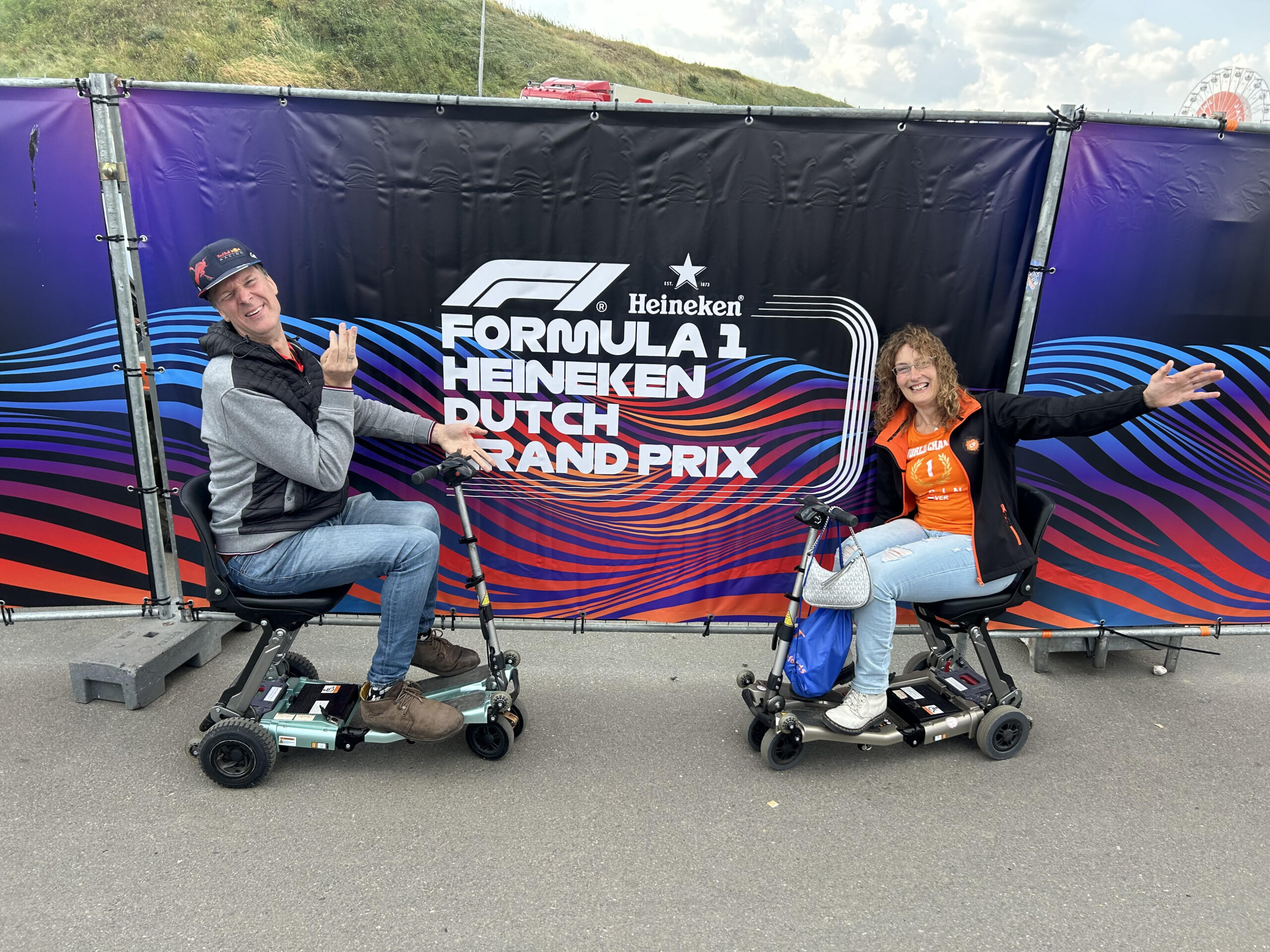 Marieke en haar man poseren op hun compacte scootmobielen voor een banner van de Formule 1.