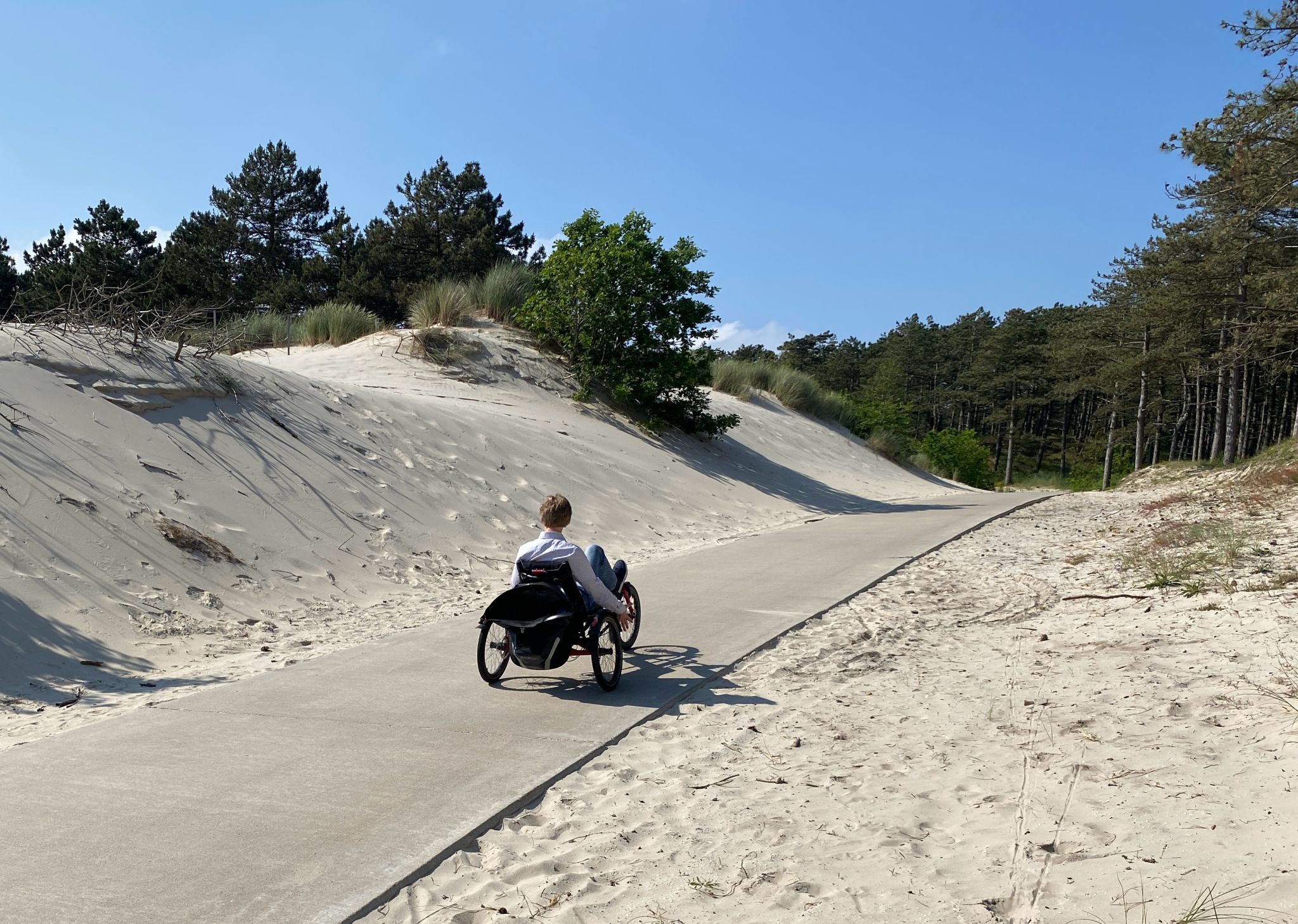je ziet mij op mijn driewielligfiets vanachter genomen. Ik fiets op asfalt langs hagelwit duinzand.
