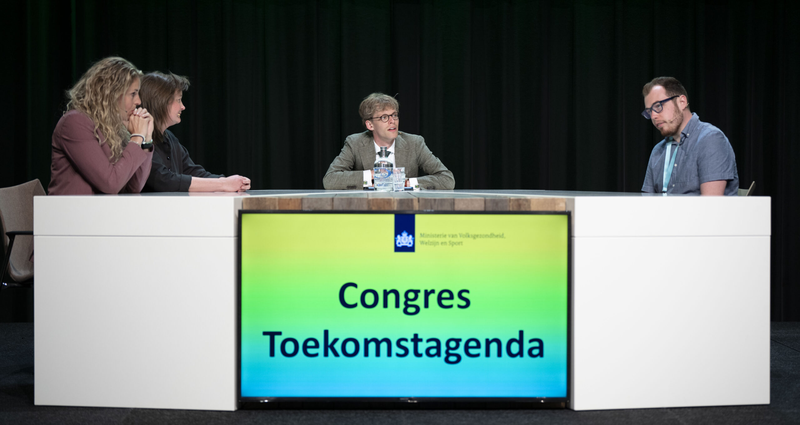 Ik zit achter een talkshowtabfel en kijk met een paar tafelgasten naar een tafelgast die praat.